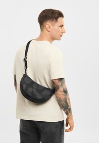 Zwarte stoffen schoudertas met een golvend patroon, gebogen vorm, verstelbare band en logo-detail, gedragen over het lichaam.