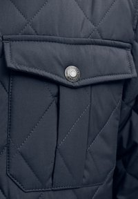 Bolsa de chaqueta acolchada azul oscuro con tapeta asegurada por un botón de presión de metal grabado "JACK & JONES EST. 1990."