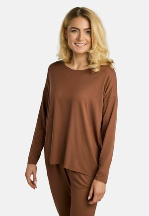 Uśmiechnięta kobieta o blond kręconych włosach, ubrana w luźną brązową bluzę z długim rękawem i pasujące spodnie, stojąca na neutralnym tle.