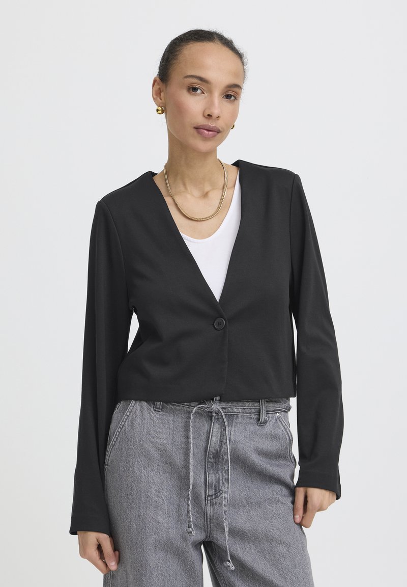 Femme portant un blazer noir à manches longues sur un haut blanc, associé à un jean gris taille haute avec une taille à cordon.