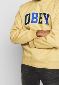 Yellow sweatshirt with "OBEY" in blue and green lettering, featuring a relaxed fit and ribbed cuffs. Worn over dark jeans.
