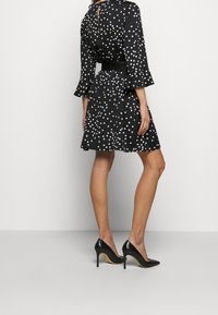 Musta mekko, jossa on valkoisia pilkkuja, lyhyet liehuvat hihat, vyötäröllä vyö ja avonainen takaosa. Yhdistetty mustiin teräväkärkisiin korkokenkiin.