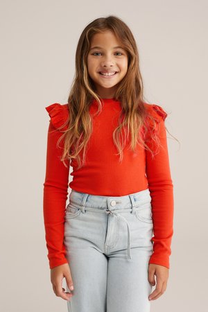 Top rosso a maniche lunghe con dettagli arricciati sulle spalle, abbinato a jeans in denim blu chiaro a vita alta con chiusura a bottoni e laccetto.