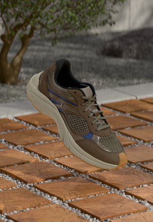 Brūni un beidžu sieta sporta apavi ar zilu akcentu un krēmkrāsas zoli, plūst virs koka un oļu flīžu grīdas tuvumā pie koka.