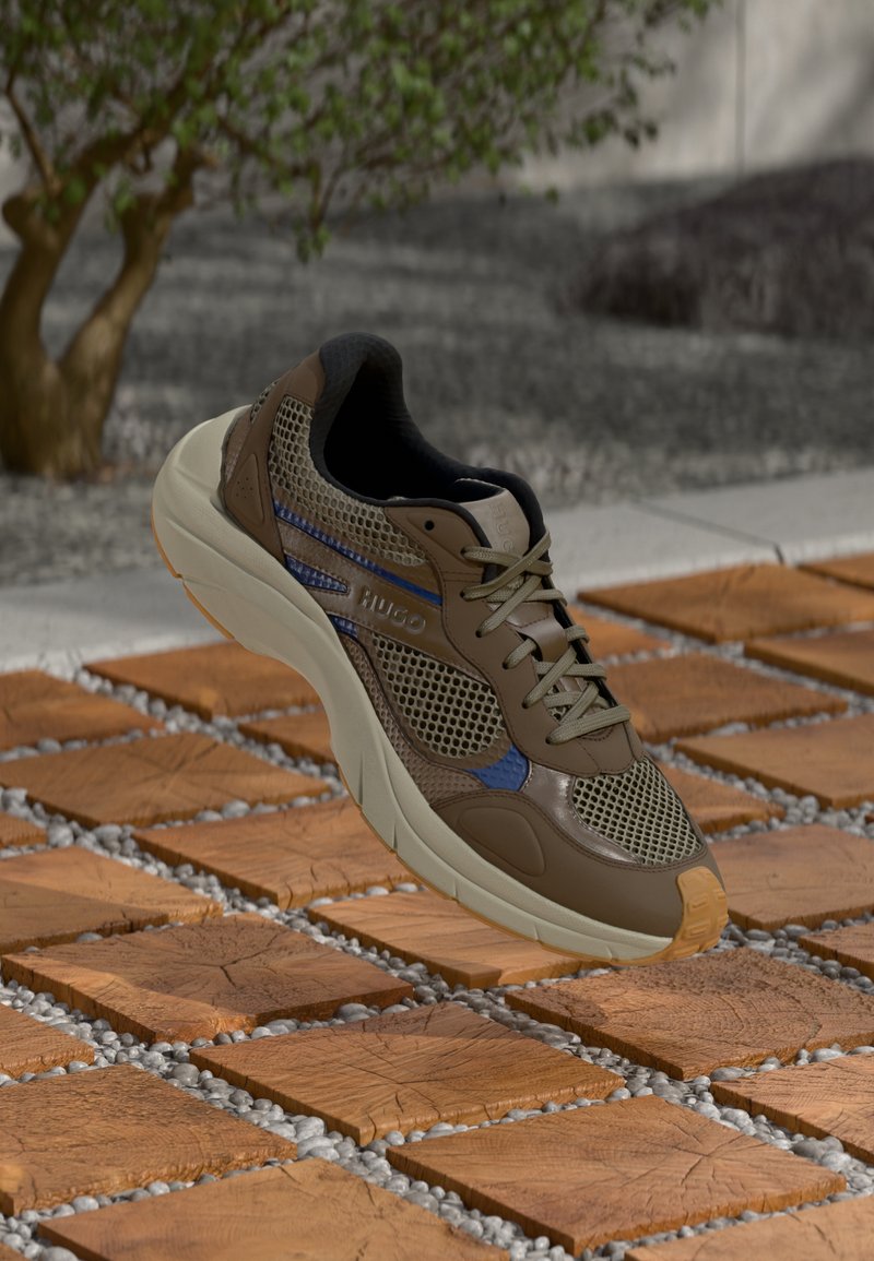 Pruun ja beež võrgust tennisekengad siniste aktsentidega ja kreemjas tald, hõljuvad puidu ja kruusa plaadi põranda kohal lähedal puule.