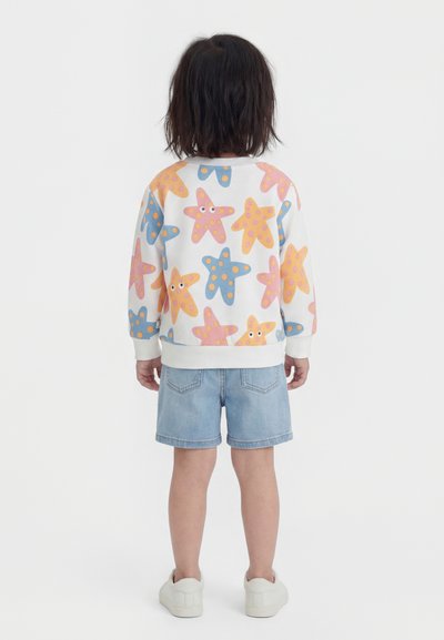 Παιδί με σκούρα μαλλιά μέχρι τους ώμους που φοράει λευκό φούτερ με πολύχρωμα σχέδια αστεριών θάλασσας, τζιν σορτς και λευκά αθλητικά, στέκεται με την πλάτη προς τα έξω.