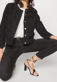 Svart denimjacka med spetsdetaljer, guldfärgade knappar, frampocket, slitna kanter, kombinerad med svarta h ögklackade sandaler med runda detaljer.