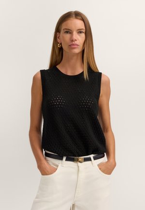 Frau mit langen, glatten blonden Haaren, trägt goldene Creolen, schwarzes ärmelloses Stricktop, weiße Hose mit schwarzem Gürtel, Hände in den Taschen.