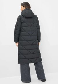 Vrouw met haar rug naar de kijker, gekleed in een lange zwarte gewatteerde jas met capuchon en een chevron-quiltpatroon, gecombineerd met een wijde, donkere spijkerbroek.