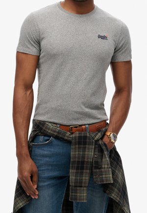 Grå t-shirt med korte ærmer lavet af bomuld, med en lille logo på venstre bryst. Kombineret med blå jeans og en ternet skjorte bundet om livet.