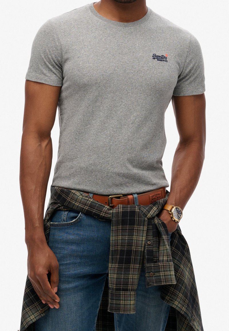 Szary, krótkorękawowy t-shirt z bawełny, z małym logo na lewej piersi. Połączony z niebieskimi dżinsami i koszulą w kratę zawiązaną wokół talii.