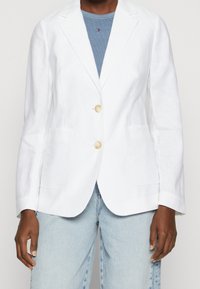Blazer de lino blanco con un corte ajustado, solapas de muesca, dos bolsillos frontales y dos grandes botones crema, combinado con una blusa azul y jeans claros.