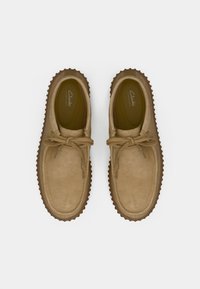 Beige velúr cipők, lekerekített orral, gumitalppal és fűzős előlappal. Jellemzője a varrási részletek és az alacsony profilú dizájn.