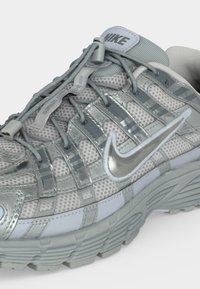 Ασημένιο και γκρι αθλητικό παπούτσι Nike με πλέγμα στο επάνω μέρος, μεταλλικό σήμα, αυλακωτή σόλα και ελαστικά κορδόνια χωρίς δέσιμο με πλαστικά κλιπ.
