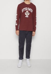 Mörkröd långärmad skjorta med texten "BITTER SWEET" och glassgrafik, med vita smiley-ikoner längs ärmarna; ihop med marinblå byxor och vita sneakers.