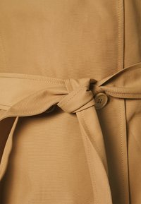 Beige trench coat s páskem, zdobení se uzlovým opaskem a knoflíkovým zapínáním, vyrobený z texturované tkaniny s viditelným šitím.