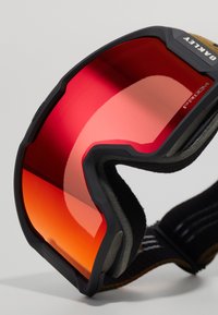 Oakley-hii suksilasit mustalla kehysillä ja punaisilla peililinssillä. Linssi on puoliympyrän muotoinen, harmaalla sisäreunuksella ja tekstuurilla varustettu hihna.