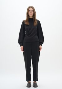 Black long-sleeve top with a high neck and gathered detailing, paired with tailored black trousers and dark slip-on shoes. Plain background.