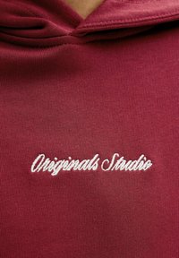 Sudadera burdeos con una textura suave y capucha. Texto bordado en blanco "Originals Studio" ubicado en el centro frontal.