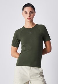 Camiseta de manga corta color verde oliva con cuello redondo y mangas raglán, que presenta un pequeño logotipo en el pecho y en una de las mangas.