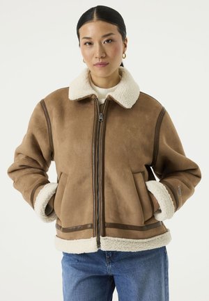 Frau trägt eine braune Lammfelljacke mit weißem Fleece-Futter, Reißverschluss vorne und blaue Jeans, Hände in den Taschen, steht vor weißem Hintergrund.