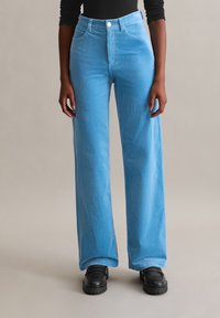 Pantalon en velours côtelé coupe large et taille haute en bleu clair. Caractérisé par une texture côtelée verticale et une fermeture avant par bouton. Porté avec des chaussures noires.