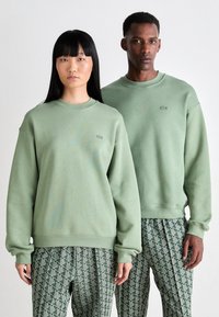 Two models wearing matching light green sweatshirts and patterned green pants, standing side by side against a plain background.