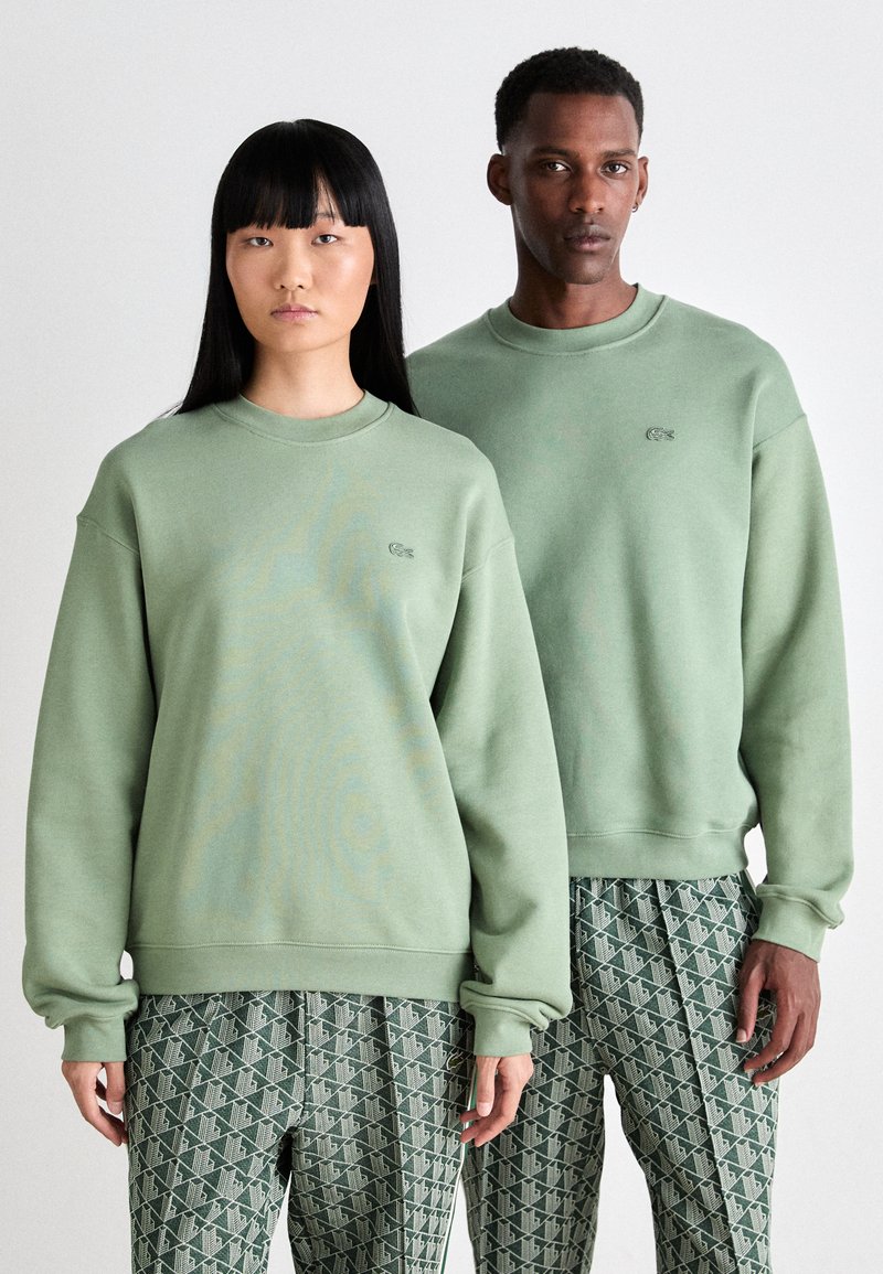 Two models wearing matching light green sweatshirts and patterned green pants, standing side by side against a plain background.