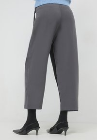 Femme portant un pantalon gris court, des collants noirs et des escarpins en vernis noir sur un fond blanc uni.