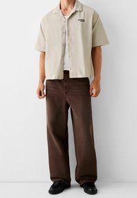 Camisa de manga corta color beige claro con botones y texto "NOMAD", combinada con pantalones marrones holgados y zapatos negros. Diseño sencillo, ajuste informal.