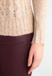 Beżowy sweter w splot kablowy z prążkowanym dołem, o luźnym kroju i teksturowanej tkaninie, w połączeniu z głęboko burgundowymi spodniami.