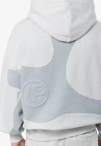 Grijze en witte hoodie met een zachte textuur, voorzien van een oversized ontwerp, groot logo-detail en contrasterende panelen op de rug.