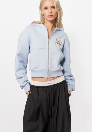 Noor naine kannab helesinist lukkudega tõmblukuga kapuutsi, millel on "LM" tikand, ja lahtisi musti pükse, millel on nähtav valge vöökoha silt "LAMANIA".