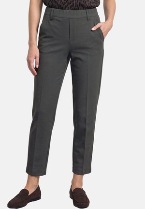 Femme portant un pantalon slim gris foncé chevilles avec poches latérales et mocassins marron foncé, debout devant un fond uni.