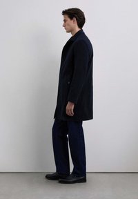 Abrigo de lana azul marino con una silueta recta, gran collar y dos botones, combinado con jeans azules y botas de cuero negras.