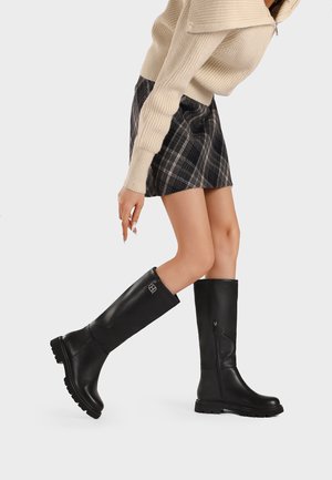 Donna che indossa un maglione beige, gonna a quadri e stivali neri al ginocchio, camminando con un piede sollevato su uno sfondo chiaro e semplice.