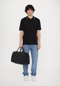 Geantă de voiaj neagră, matlasată, cu formă rotunjită, mânere duale și închidere cu fermoar. Modelul poartă un polo negru și blugi deschiși, cu adidași albi.