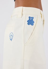 Un pantalon de couleur crème présente des graphiques brodés bleus : un ours et un smiley avec un sourire dégoulinant. Le tissu a une apparence lisse et texturée.