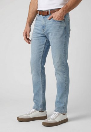 Mann trägt hellblaue Jeans im geraden Schnitt, braunen Ledergürtel, weiße Sneaker und ein weißes Hemd, mit einer Hand in der Tasche.