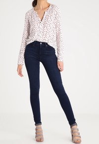 Vit långärmad blus med ett litet mönster av rosa och svarta fåglar, tillsammans med mörkblå skinny jeans och grå klackskor med remmar.