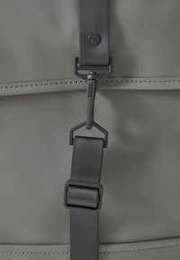 Dettaglio di uno zaino impermeabile grigio con finitura opaca, cinturino regolabile e logo sulla tracolla, che evidenzia linee pulite e cuciture.