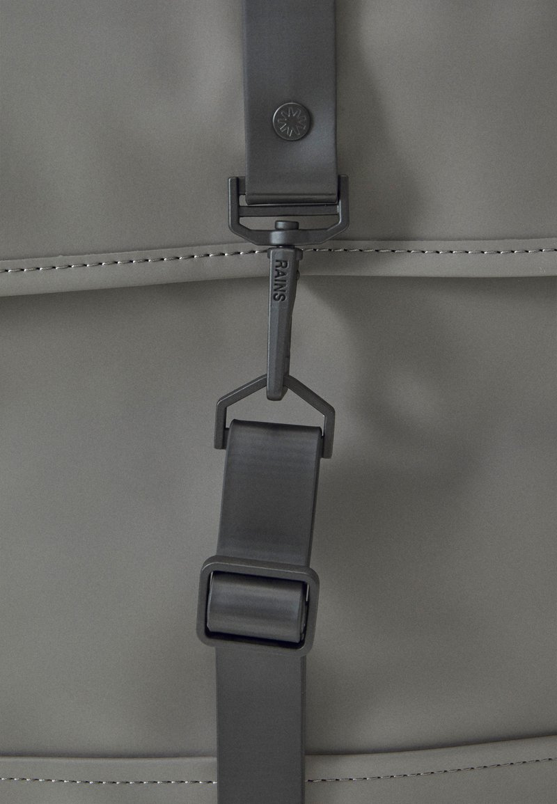 Détail d'un sac à dos imperméable gris présentant une finition mate, une boucle ajustable et un logo sur la sangle, mettant en avant des lignes épurées et des coutures.