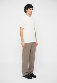 Άνδρας που στέκεται φορώντας λευκό κοντομάνικο πόλο μπλούζα, καφέ φαρδιά παντελόνια και μαύρα δερμάτινα παπούτσια, μπροστά σε απλό φόντο.