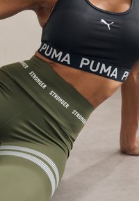 Gros plan sur une personne portant un soutien-gorge de sport noir Puma et un legging vert olive avec le texte "STRONGER", posant au sol pendant un entraînement.