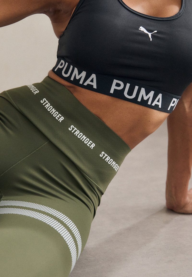 Gros plan sur une personne portant un soutien-gorge de sport noir Puma et un legging vert olive avec le texte "STRONGER", posant au sol pendant un entraînement.