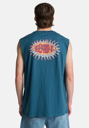 Uomo con maglietta senza maniche color teal con logo stilizzato di sole arancione e rosso e testo sulla parte superiore della schiena, abbinata a pantaloncini di jeans azzurri.