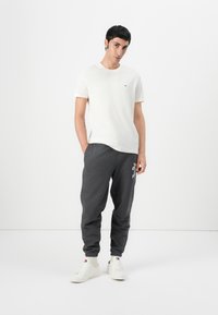T-shirt en coton blanc avec un petit logo sur la poitrine, associée à un pantalon de survêtement gris foncé avec du texte imprimé, et des baskets blanches.