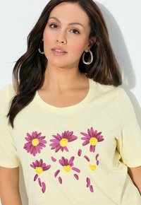 �Żółta koszulka z krótkim rękawem z kwiatowym wzorem z fioletowymi kwiatami i żółtymi środkami, wykonana z miękkiego, gładkiego materiału.