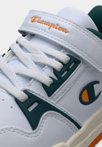 Λευκό αθλητικό παπούτσι με υφή στην επάνω πλευρά, πράσινες λεπτομέρειες και πορτοκαλί λογότυπο «Champion». Διαθέτει σχέδιο με κορδόνια και επένδυση στο λουρί για ασφαλή εφαρμογή.