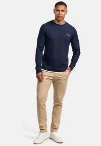 Marinebluse med struktur og lange ærmer samt rund hals, parret med lyse beige bukser og hvide sneakers, med diskret logo detalje på brystet.